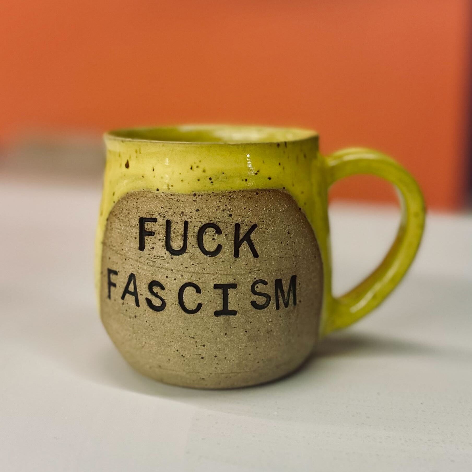 Mug with 'FUCK FASCISM' text on a white surface with an orange background